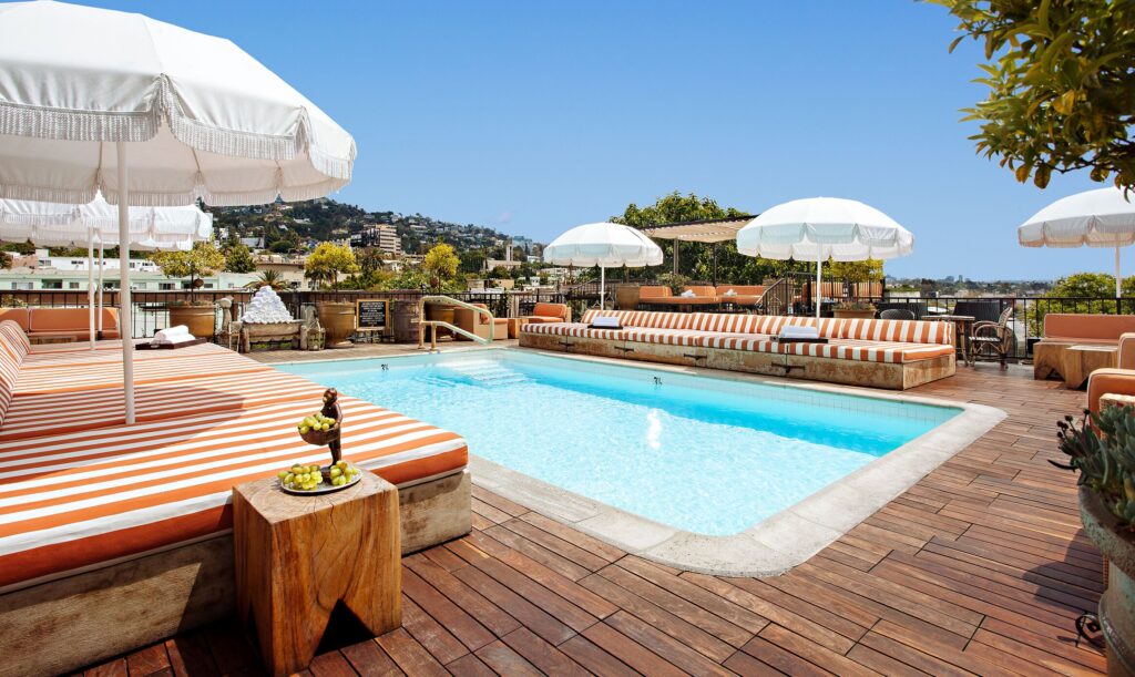 The rooftop wooden pool deck at Petit Ermitage, complete with bohemian white umbrellas and striped lounge seats, on a sunny day in West Hollywood.