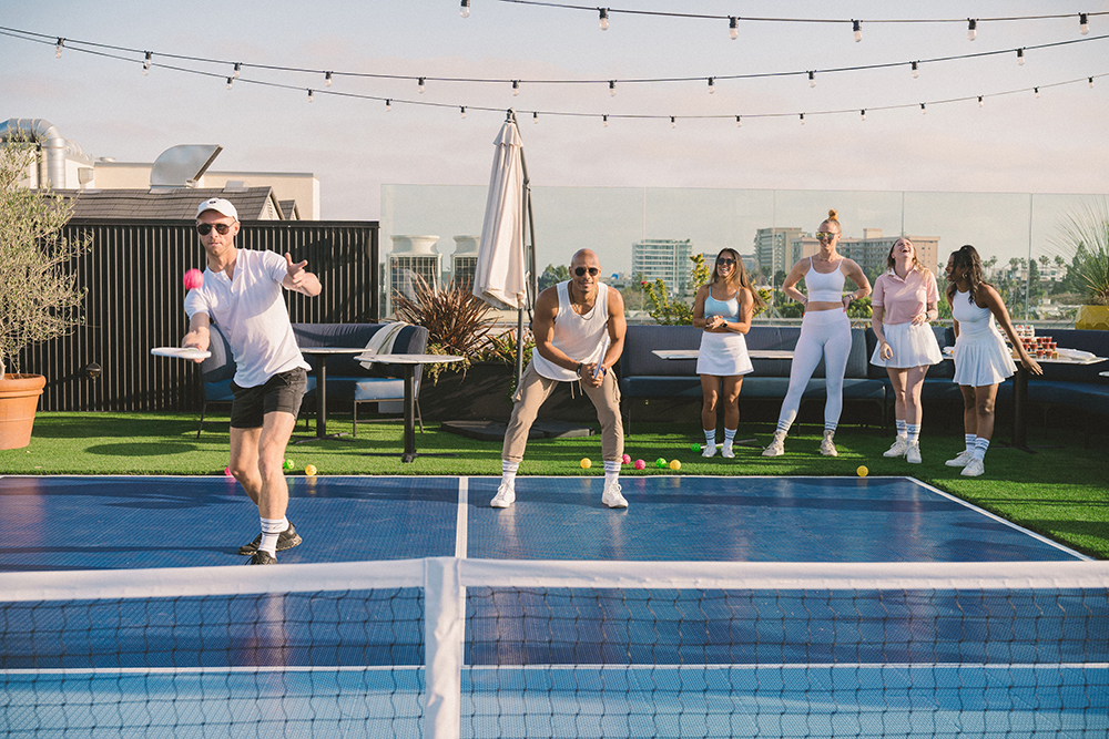 Four women laughing while watching their two male friends play pickleball at the rooftop court at Kimpton La Peer Hotel.