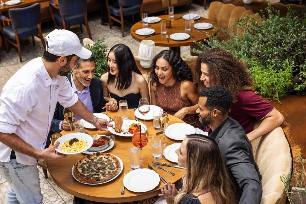 A chef presents plates of food to a large table of excited guests in the outdoor patio of Lavo. West Hollywood, California.